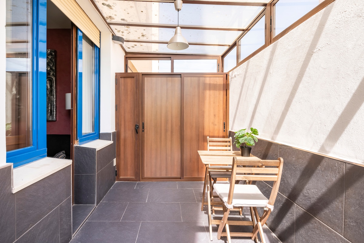 An enclosed terrace is presented with natural light filtering through glass panels above. A small table with two chairs is located on a tiled floor, alongside a wooden cabinet and a potted plant. Blue-framed windows add a pop of color to the space.