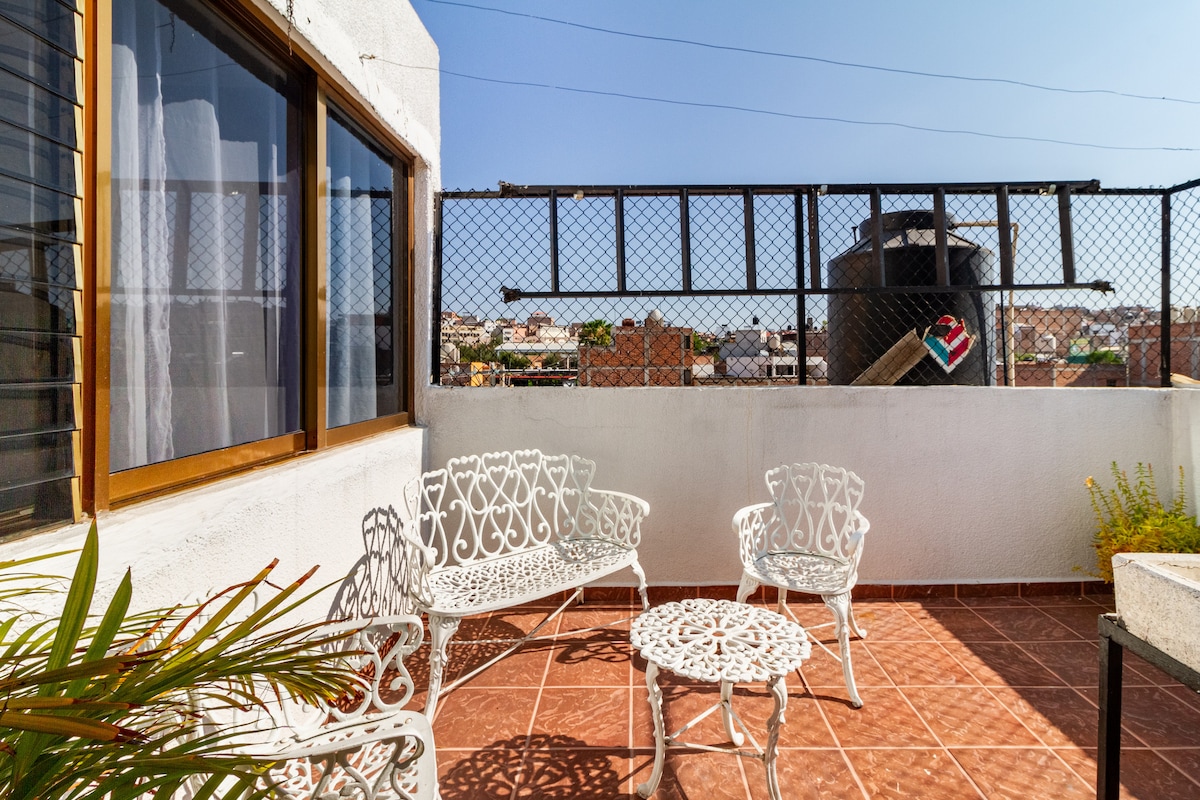 A small terrace is adorned with white metal furniture, including a round table surrounded by four chairs. Terracotta tiles create a warm ambiance. In the background, a fenced area offers views of the city skyline, with clear skies overhead.