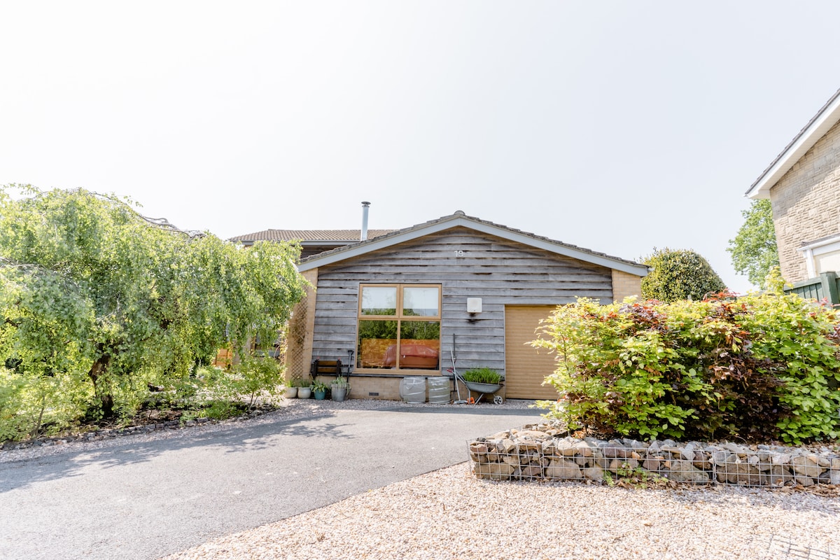 The exterior of the family home features a wooden facade with a large window, providing visible natural light. A gravel driveway leads to a garage, and landscaped greenery surrounds the property, enhancing the welcoming atmosphere of the entrance area.