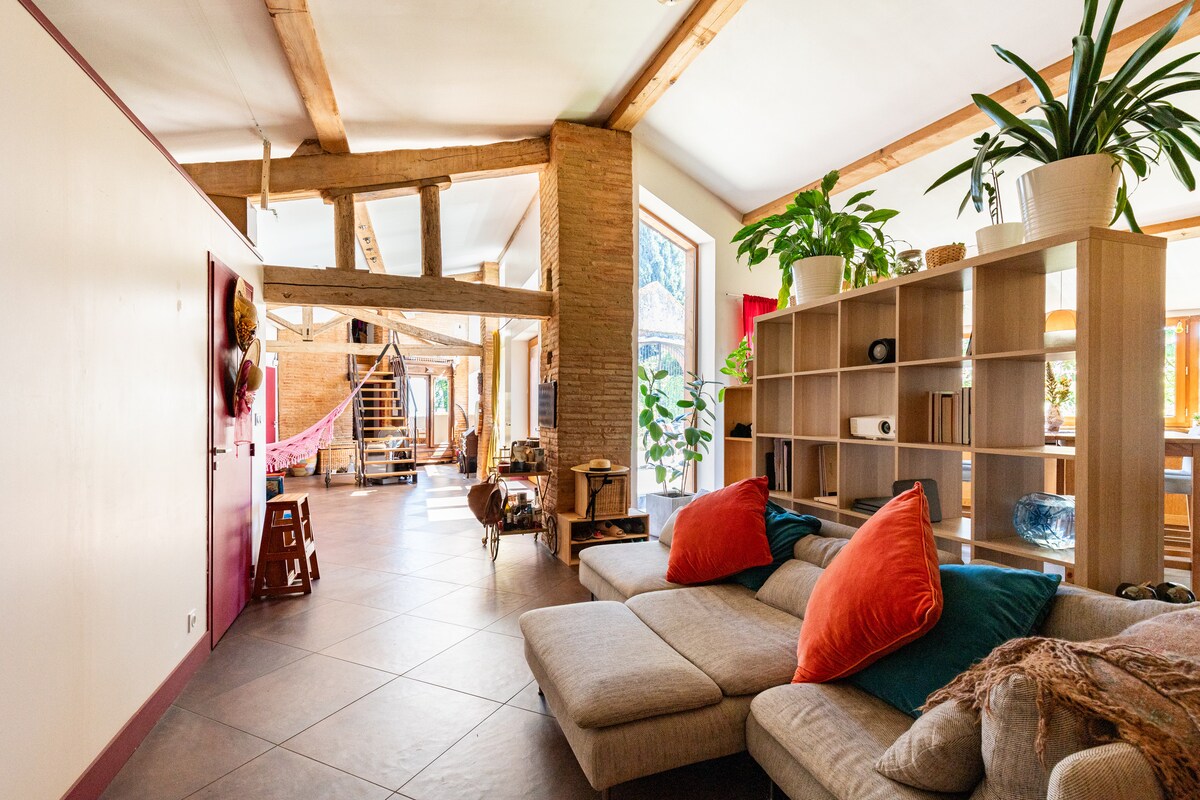 A spacious living area features a light-hued sectional sofa adorned with two colorful throw pillows. Natural light filters through large windows, illuminating the space filled with greenery and modern decor. Exposed wooden beams add character to the high ceiling, enhancing the openness of the room.