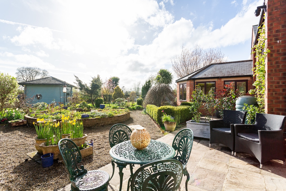 A private patio area is featured, furnished with an intricately designed table and chairs. Lush greenery surrounds the space, with a variety of blooms and garden beds visible. Natural light streams in, enhancing the tranquil ambiance of the outdoor setting.