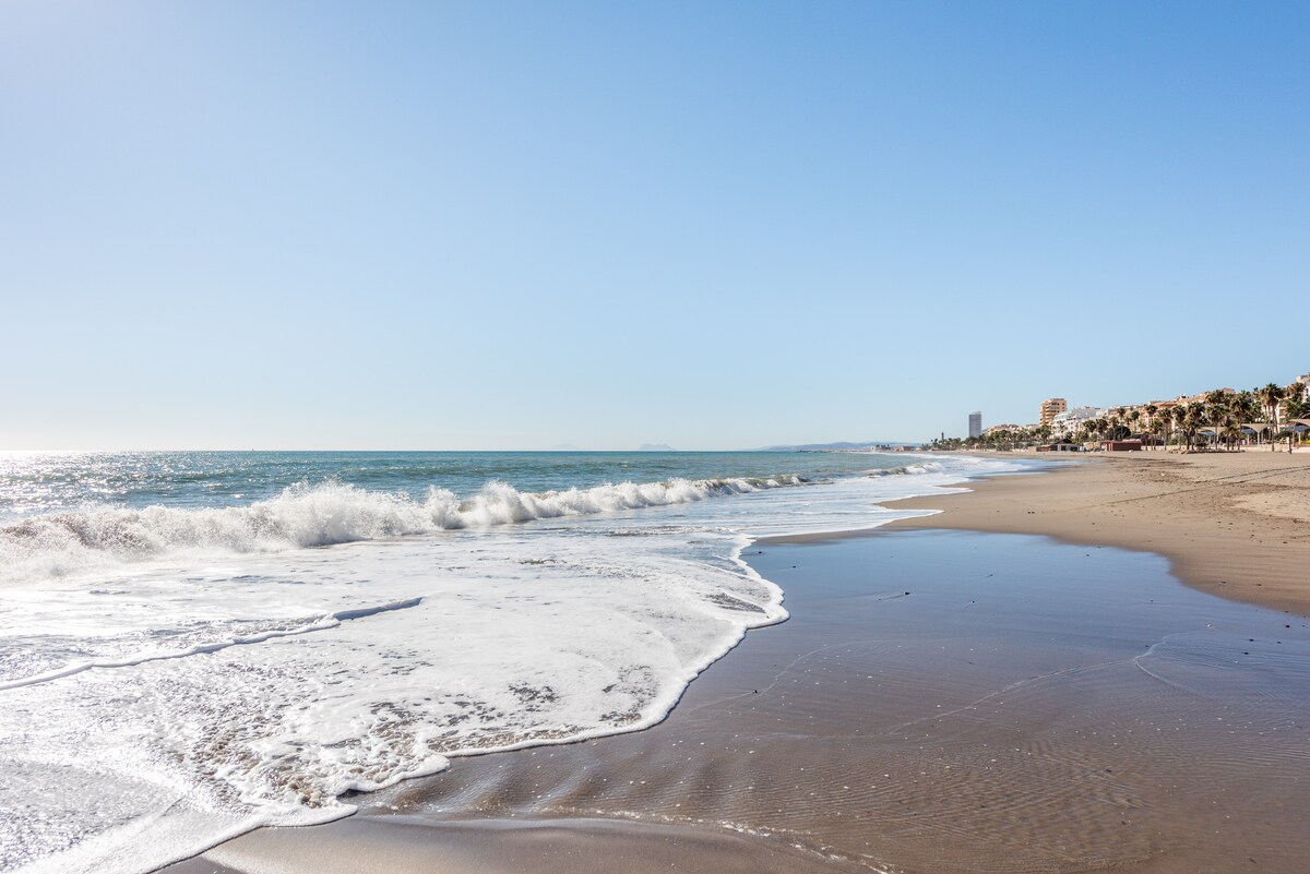 A serene beachfront scene captures gentle waves lapping at the shore under a clear blue sky. The wet sand reflects the sunlight, creating a shimmering effect. The distant coastline is visible, revealing a mix of buildings against the backdrop of the horizon.