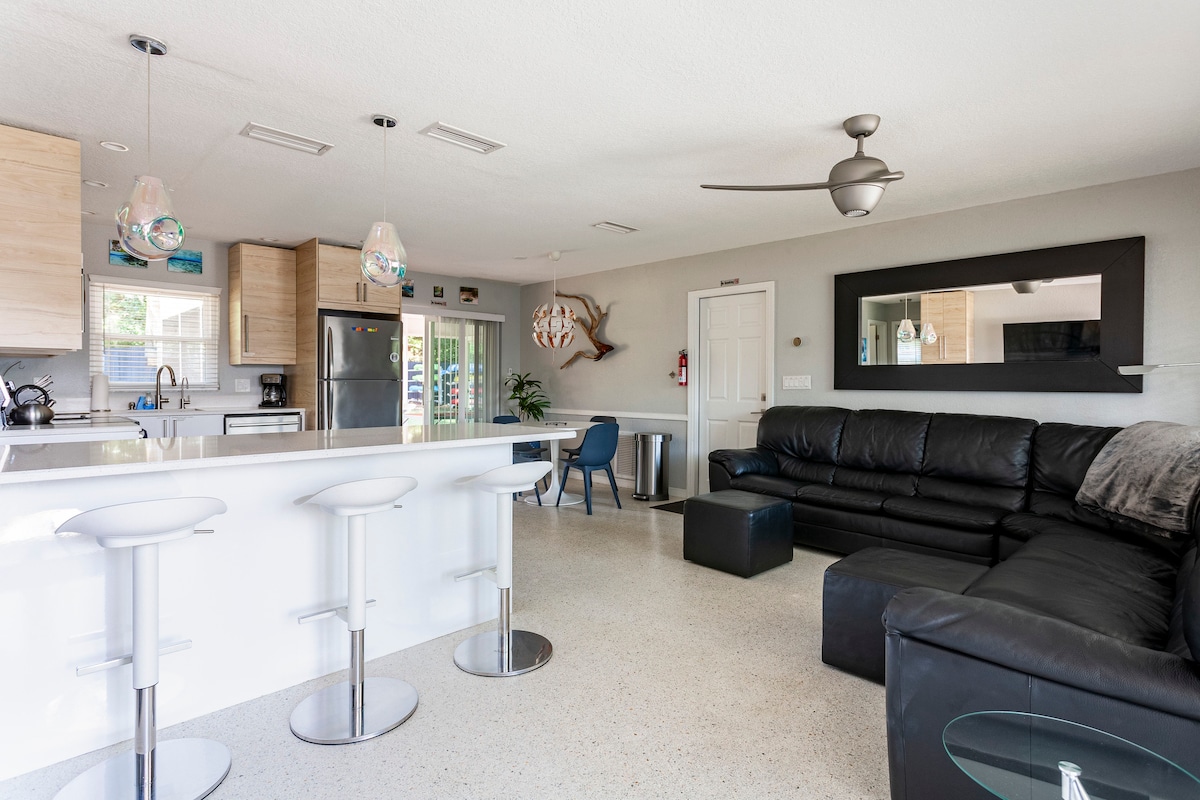 An open-concept living area is visible, featuring a modern kitchen with sleek cabinetry and stainless-steel appliances. A black leather sectional sofa provides ample seating, complemented by stylish bar stools at the counter. Natural light illuminates the space through sliding glass doors.