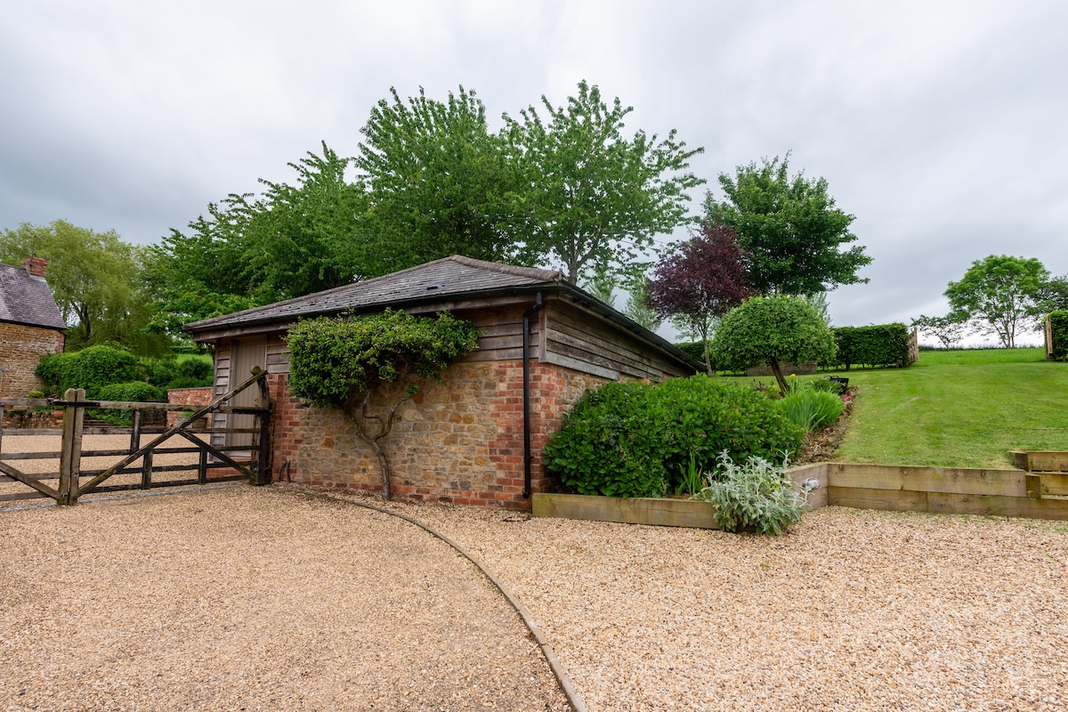 The exterior of The Old Stable features a combination of stone and wood finishes, surrounded by landscaped greenery. A gated entrance leads to a gravel pathway, while a manicured hedge and trees provide a natural backdrop, creating a calm and inviting setting.