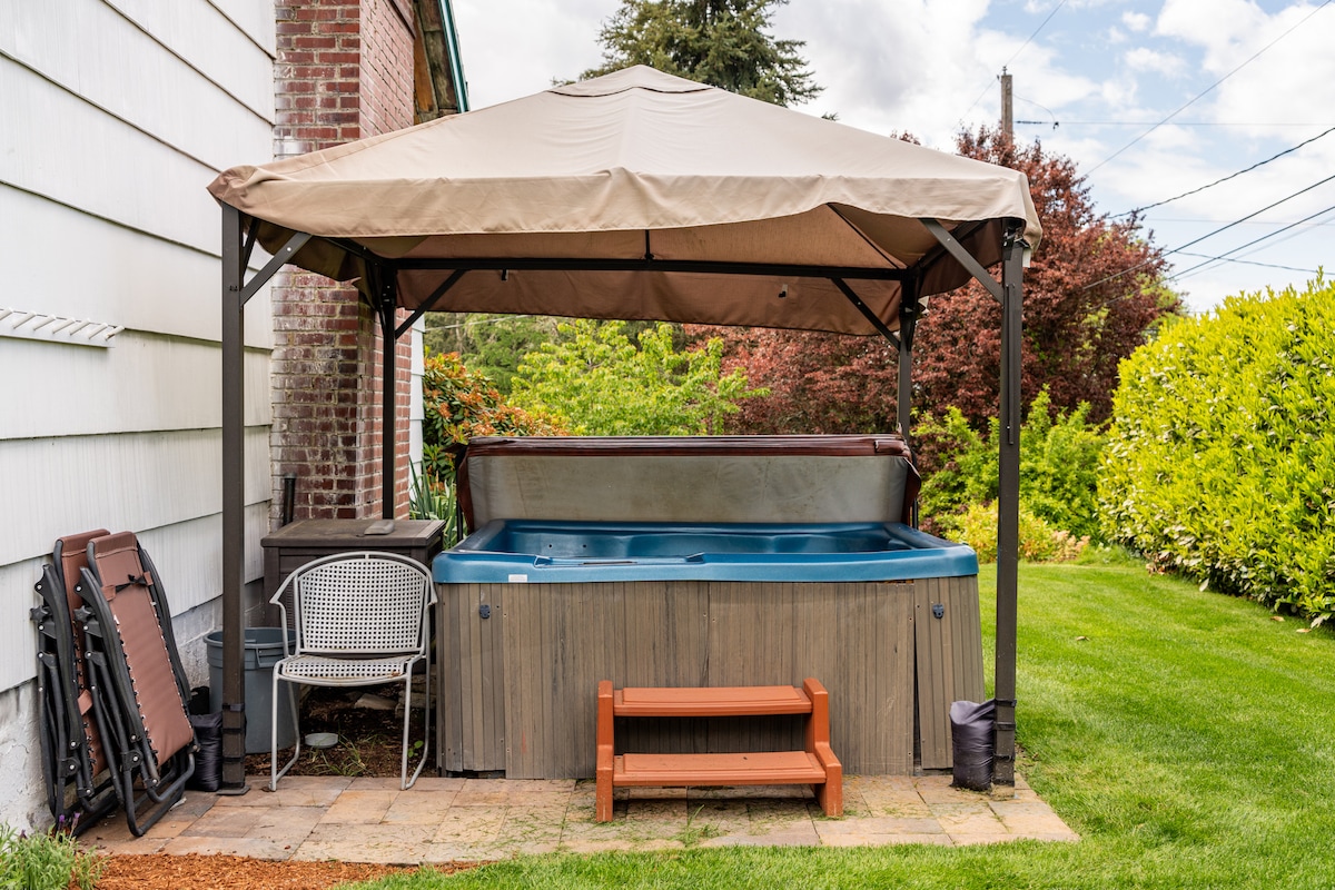 A hot tub is sheltered under a beige canopy, surrounded by a green lawn. A set of folding chairs is neatly stacked nearby, and a wooden bench sits in front of the hot tub. Lush greenery can be seen in the background, creating a serene outdoor space.