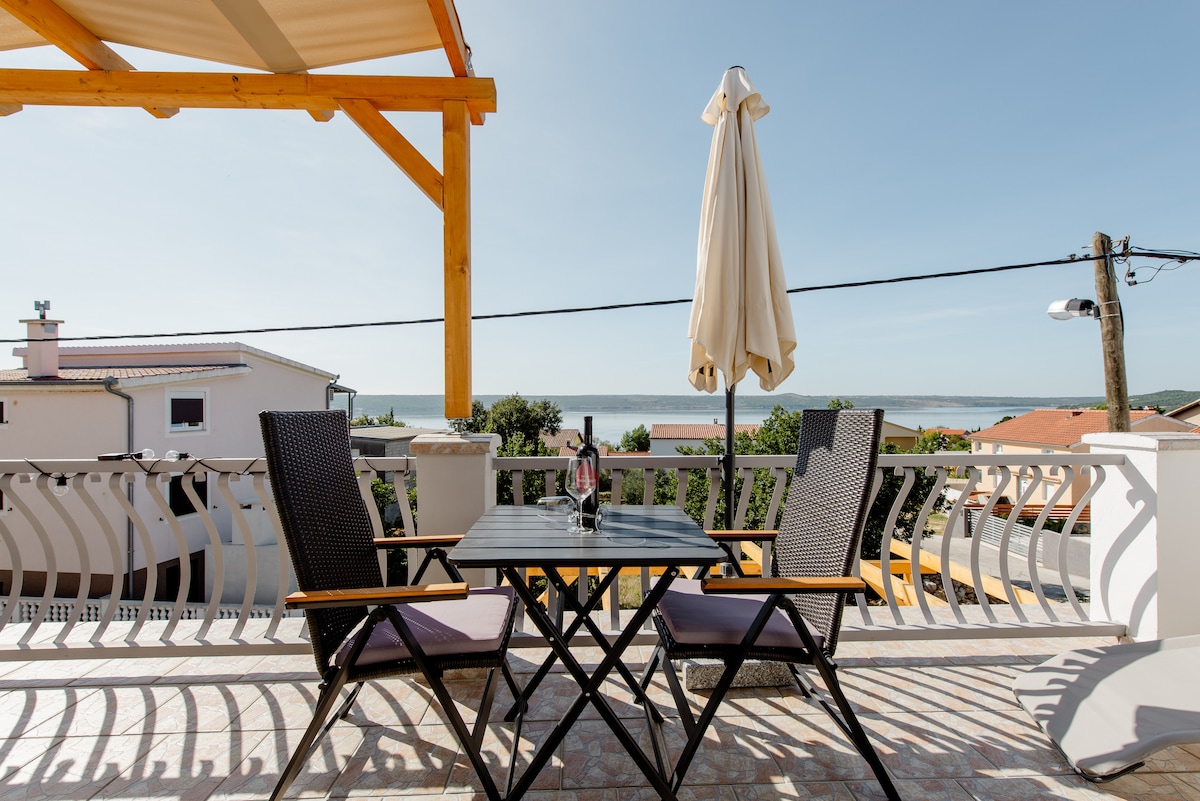 A spacious terrace is presented, featuring a table and four chairs, accompanied by a standalone umbrella. A scenic view of the surrounding area and the sea can be seen in the background, framed by a wooden pergola, enhancing the outdoor dining experience.