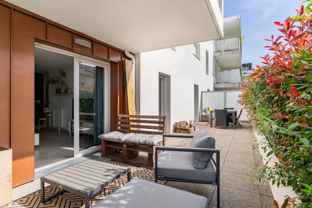 A spacious terrace is adorned with comfortable seating options and a coffee table. Lush greenery frames the area, while light-colored walls of the residence create a serene environment. Sliding glass doors provide a seamless connection to the indoor living space.