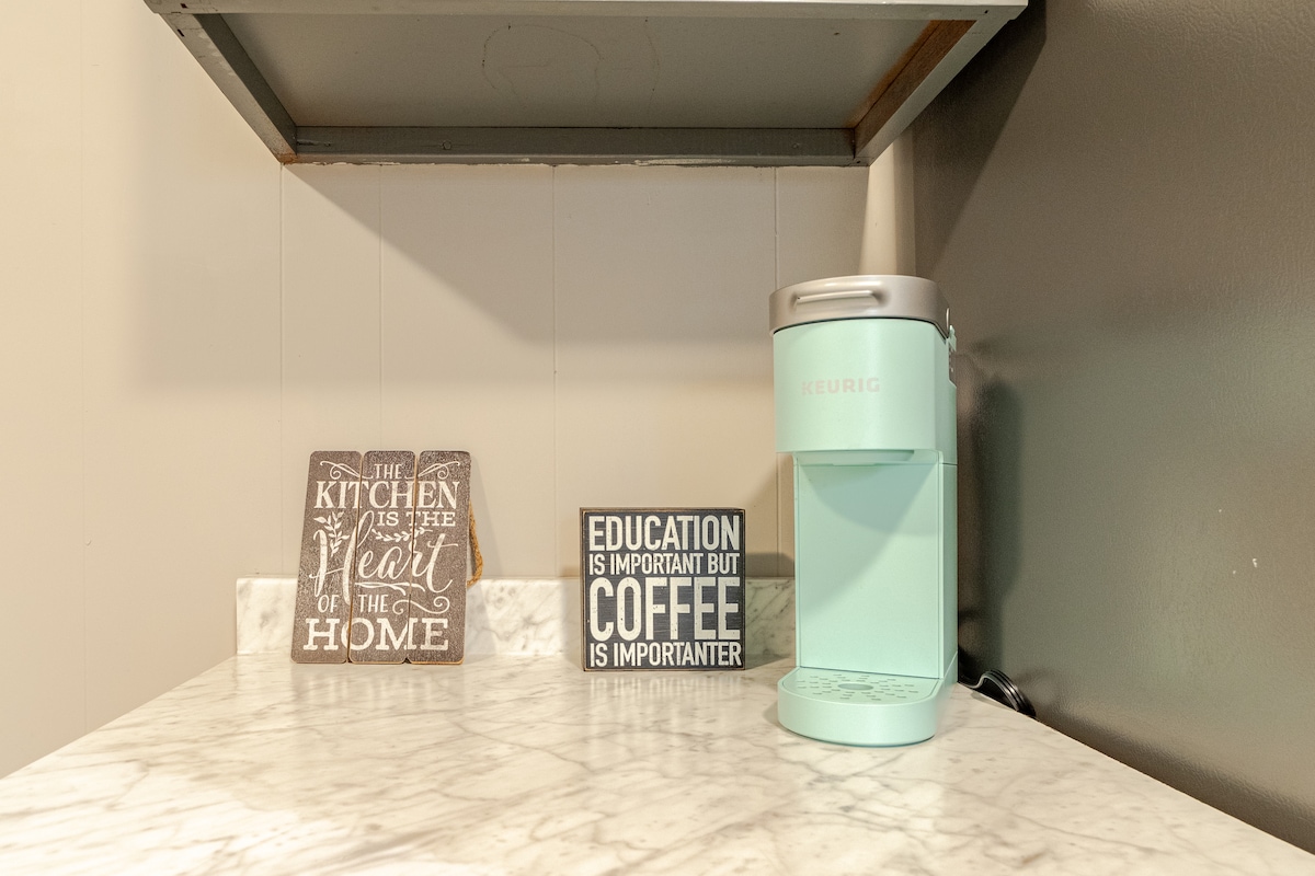 A modern countertop is featured with a light marble finish, showcasing a teal coffee maker beside two decorative signs. One sign highlights the kitchen as the heart of the home, while the other emphasizes the importance of education over coffee.