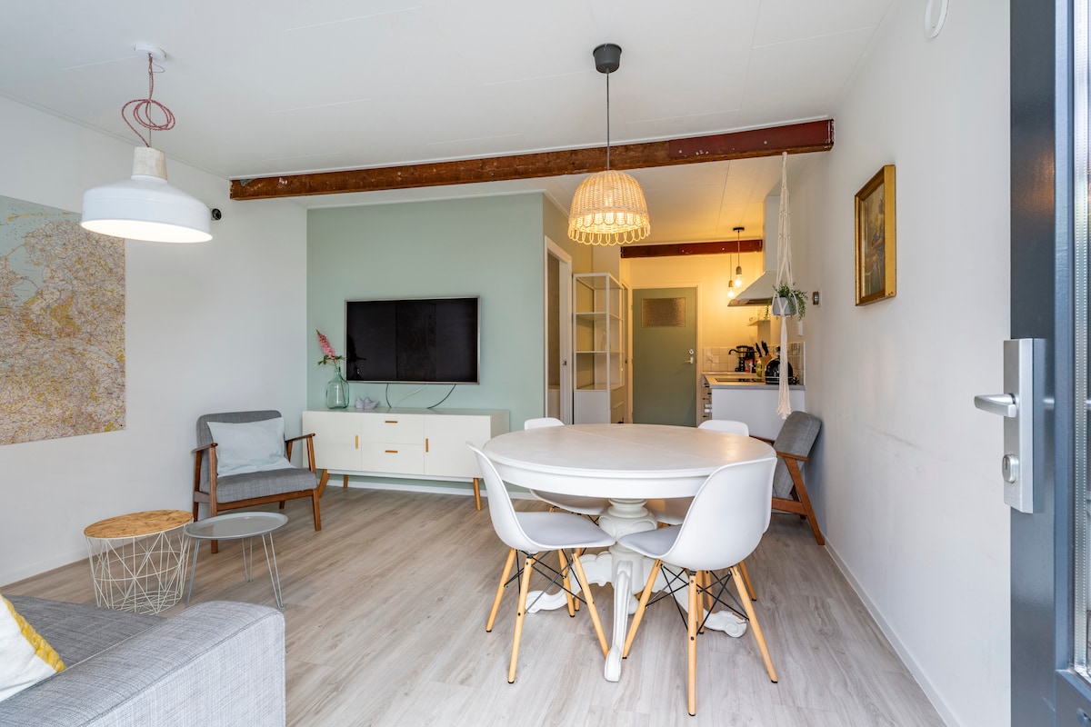 A light and airy living area is showcased, featuring a round dining table surrounded by white chairs. A comfortable seating area includes two armchairs and a modern coffee table. A television is mounted on the wall, and a kitchen can be seen in the background.