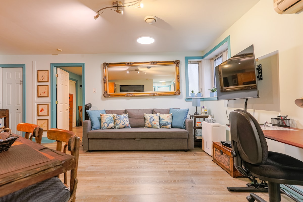 A living area is displayed featuring a comfortable gray sofa adorned with colorful accent pillows. A decorative mirror hangs above the sofa, while a wall-mounted TV is visible. A workspace with a chair and wooden dining table occupy the space, complemented by natural light from nearby windows.