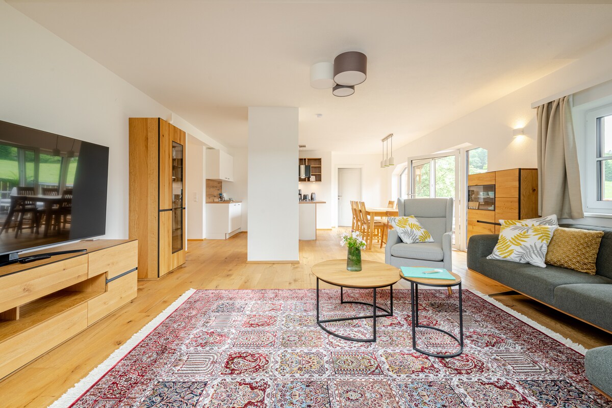 A spacious living area features a large television mounted on a wooden unit, complemented by modern seating. A round coffee table sits on a patterned rug, while a kitchen area is visible in the background. Natural light streams through the windows, creating an open atmosphere.
