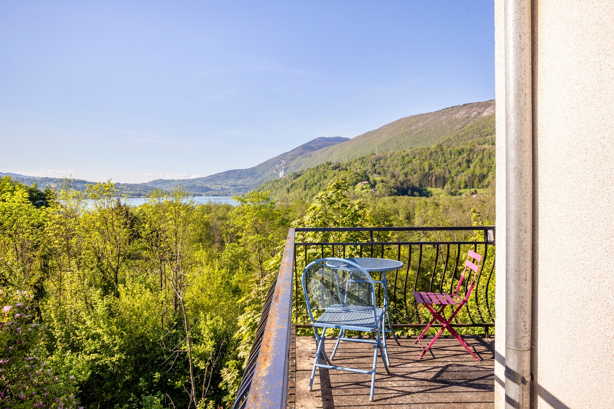 A balcony overlooks a serene landscape of greenery and hills, with the lake visible in the distance. A round glass table is accompanied by colorful chairs, creating an inviting space to enjoy the view while surrounded by nature.