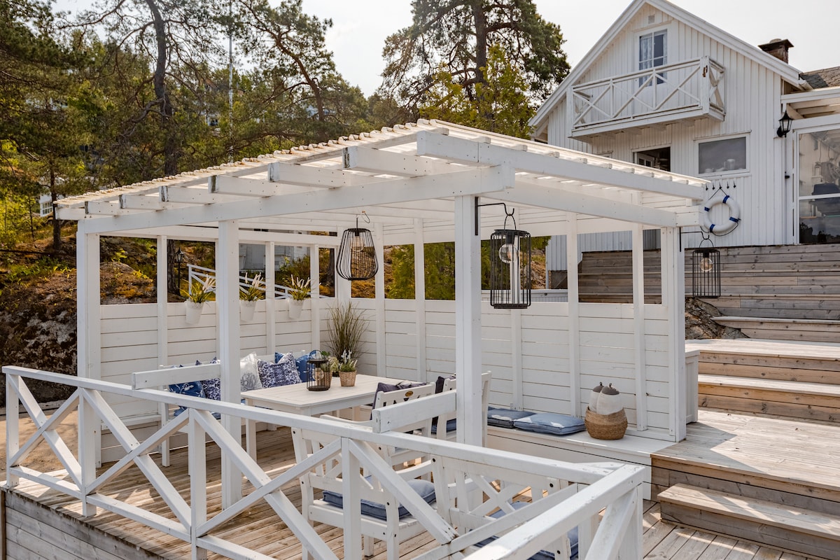 An outdoor terrace features a white wooden structure providing shade. A table is surrounded by seating, enhancing the space for relaxation or dining. Lanterns hang from the ceiling, and potted plants add subtle greenery. Steps lead up to a light-colored building in the background.