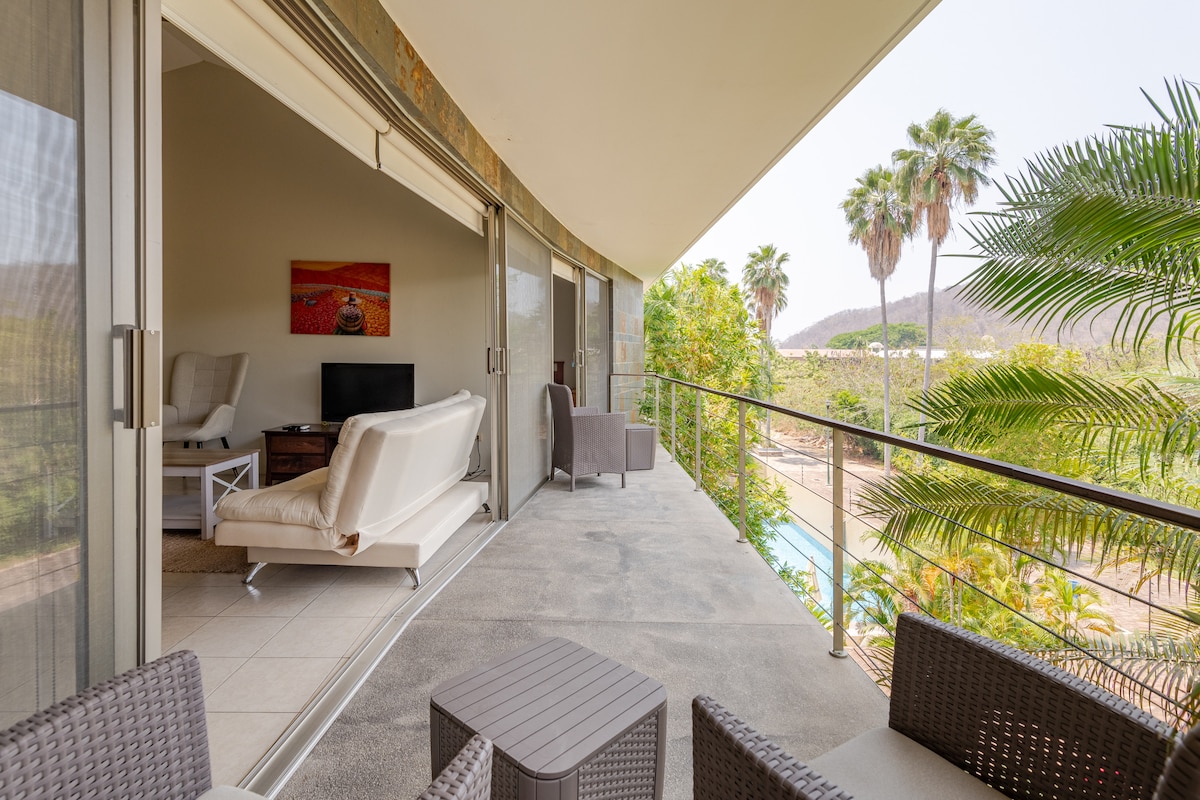 An expansive balcony is shown, featuring a set of comfortable seating arrangements and a small table. Lush palm trees can be seen beyond the railing, along with a clear view of the surrounding landscape and a glimpse of the pool area.