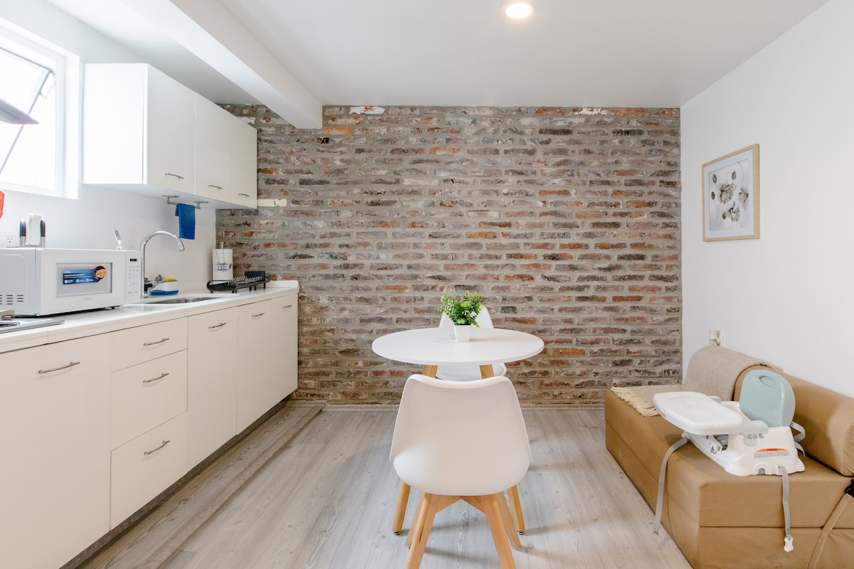 A modern kitchen area features a combination of white cabinetry and exposed brick walls, creating a stylish contrast. A round dining table is surrounded by sleek chairs, with a cozy sofa nearby. Natural light enters through a window, enhancing the space's inviting ambiance.