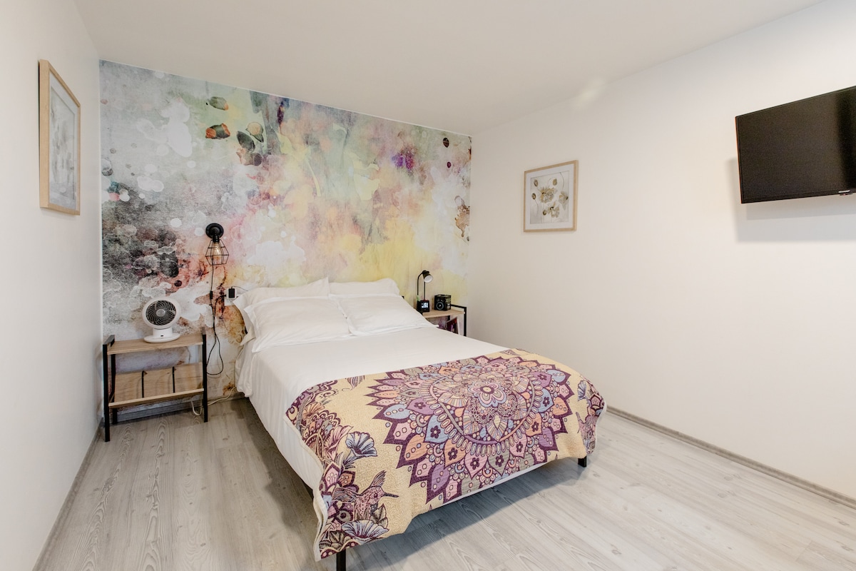 The room features a spacious bed adorned with a vibrant patterned blanket. A large mural with soft colors serves as a backdrop. A flat-screen TV is mounted on the wall, with a small nightstand beside the bed. Natural light enhances the light wooden flooring.