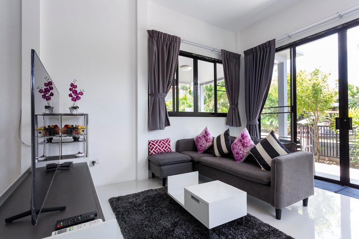 A modern living area features a spacious gray sectional sofa adorned with vibrant purple and white throw pillows. Large windows are framed by dark curtains, allowing natural light to brighten the space. A sleek TV stand displays a collection of decorative items, enhancing the contemporary atmosphere.