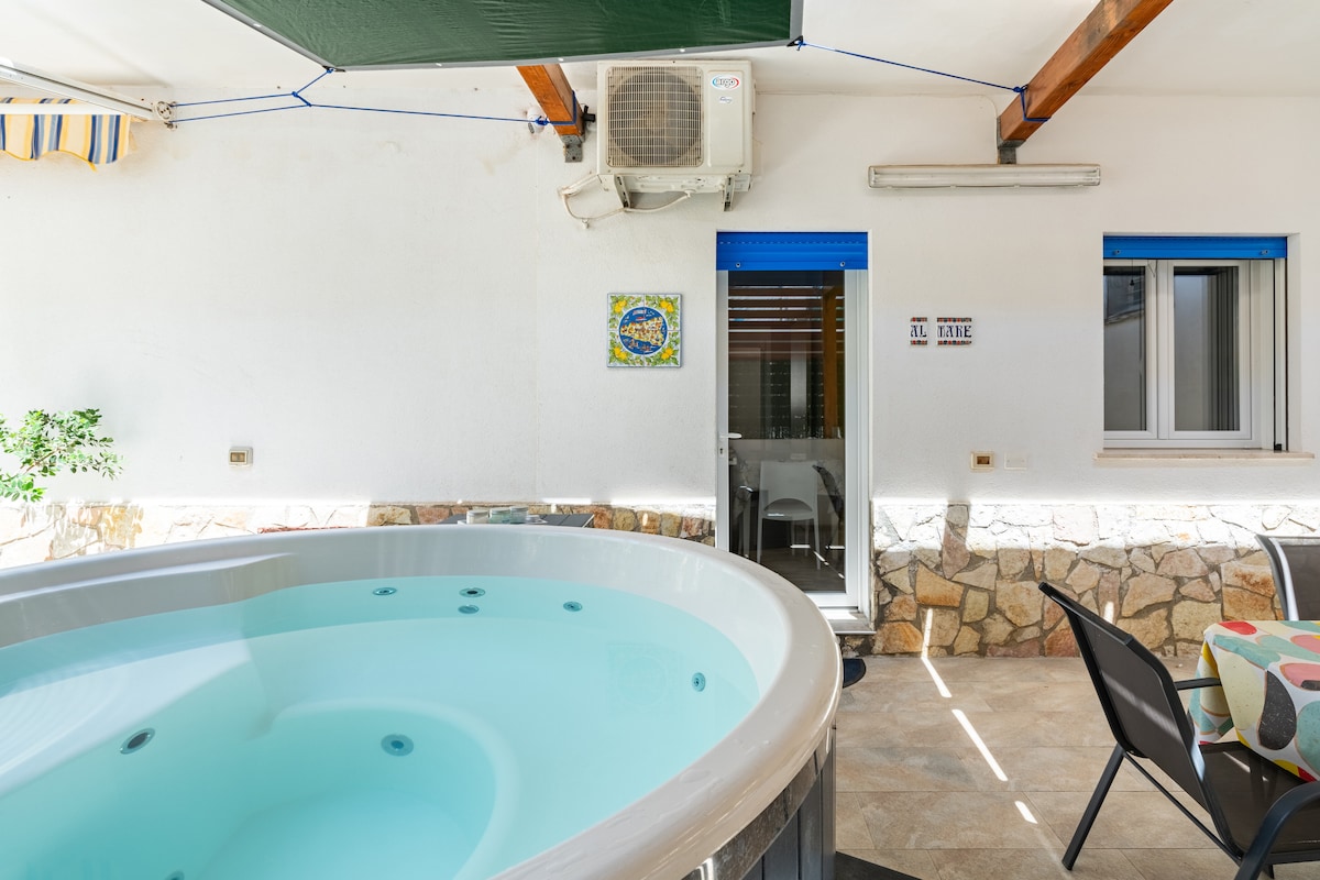 An outdoor area features a heated whirlpool tub, surrounded by a stony wall. A small patio table and chairs are visible, along with an air conditioning unit mounted on the wall above. A doorway leads into the interior space, with additional windows offering natural light.