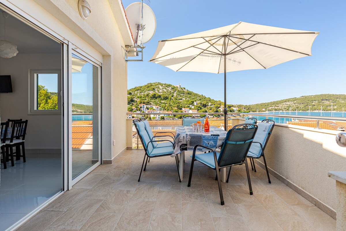A private terrace is showcased with a dining table set under a large umbrella. Comfortable chairs surround the table, providing a relaxing space with views of the surrounding hills and coastline.