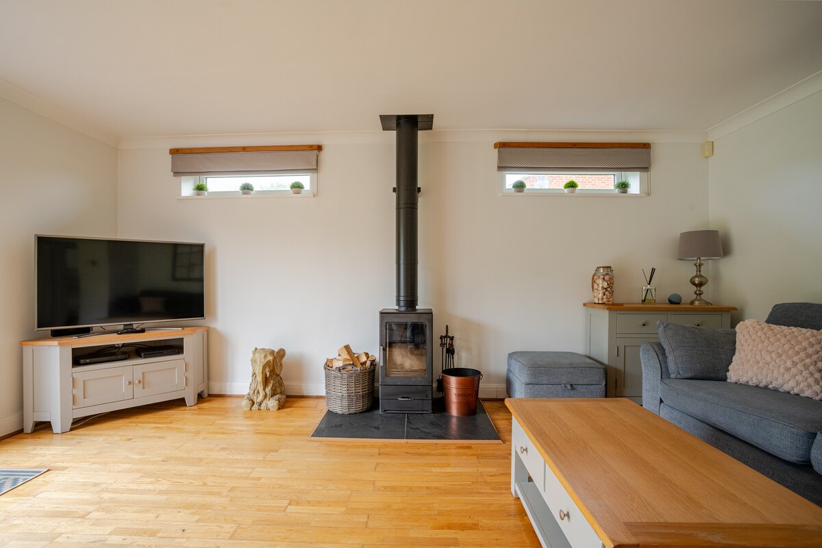 A welcoming living room features light wood flooring and soft, neutral walls. A log burner stands against one wall, accompanied by a television on a media unit. Two comfortable sofas are positioned for relaxation, with a coffee table centrally placed for convenience.