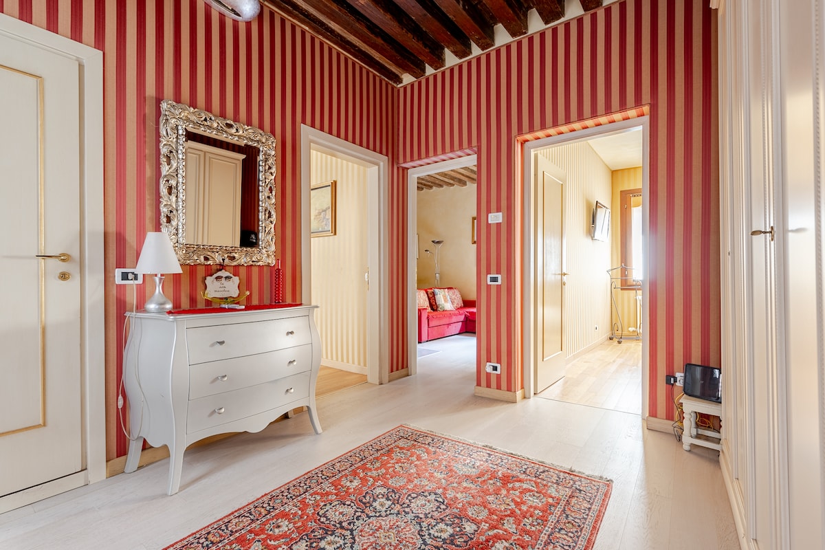 A striped hallway features warm red and beige tones, showcasing a decorative mirror above a light-colored dresser. A patterned rug is placed centrally, enhancing the space. Doorways lead to adjoining rooms, while light enters through windows at the far end.