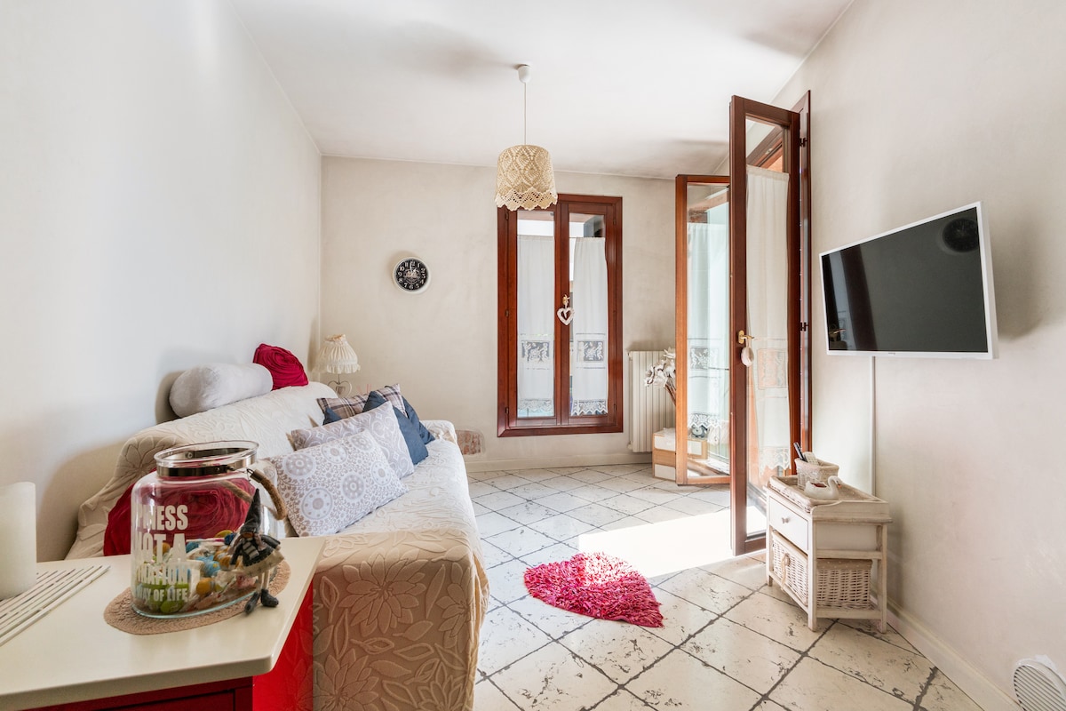 The living area features a comfortable sofa adorned with decorative cushions and a small red rug on the tiled floor. Large glass doors open to a balcony, allowing natural light to illuminate the space. A wall-mounted TV and a small side table complete the inviting setting.