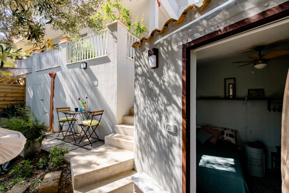 An independent entrance is visible, leading to a cozy space surrounded by greenery. A small outdoor seating area with a table and chairs offers a peaceful spot for relaxation. Natural light filters through the trees, casting dappled shadows on the steps.