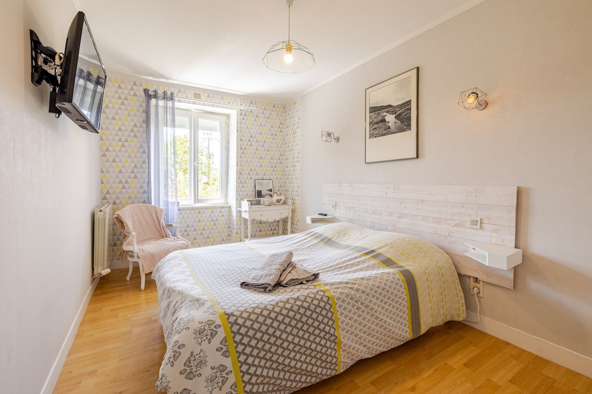 A spacious bedroom features a large bed with a patterned duvet cover. A desk and a chair are positioned near a window, allowing natural light to fill the room. Decorative lighting fixtures and a wall-mounted television add a modern touch to the inviting space.