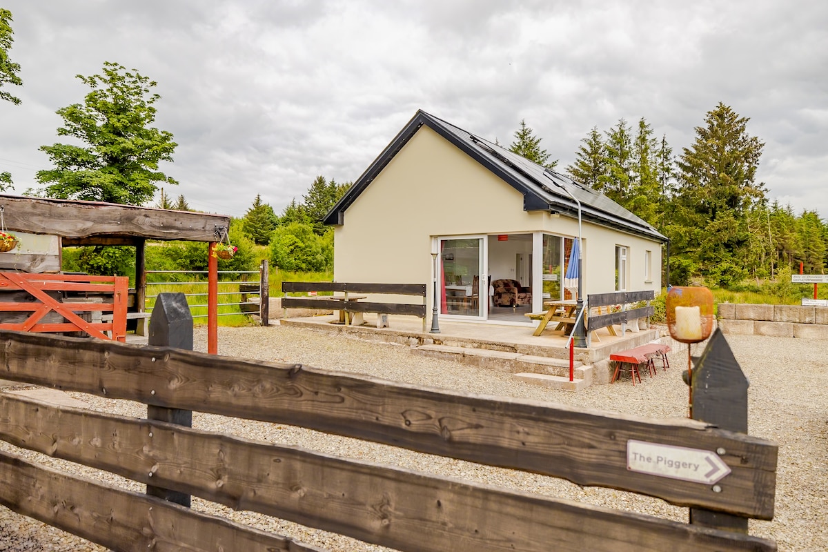 The renovated cottage features a light-colored exterior with large glass doors offering views into an open area. Surrounding wooden fencing provides an inviting entrance, complemented by trees and greenery in the background.