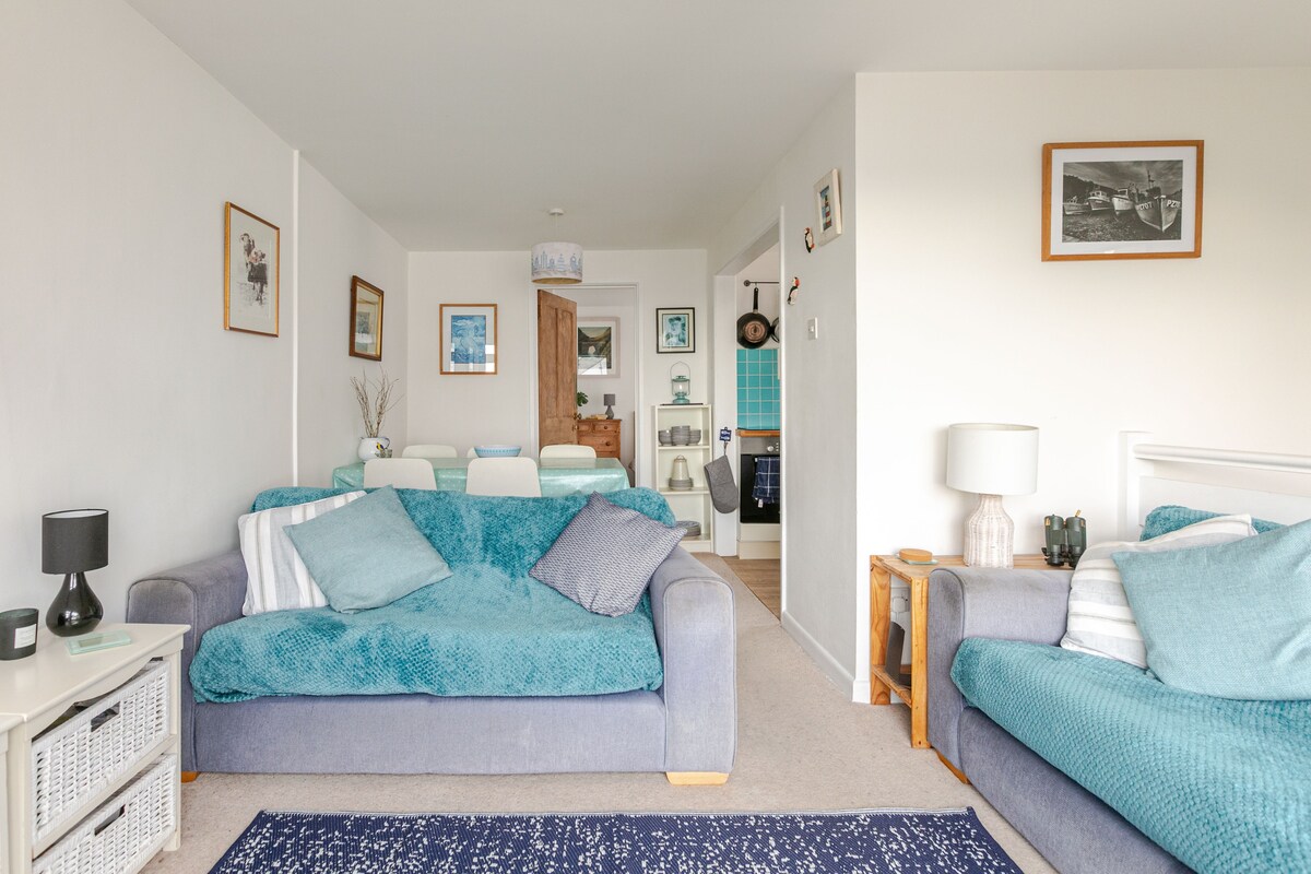 A light-filled lounge area features two comfortable blue sofas adorned with textured throws. A patterned area rug lies on the carpeted floor. Pictures and decor are displayed on the walls, creating a relaxed atmosphere that leads into a dining and kitchen space in the background.