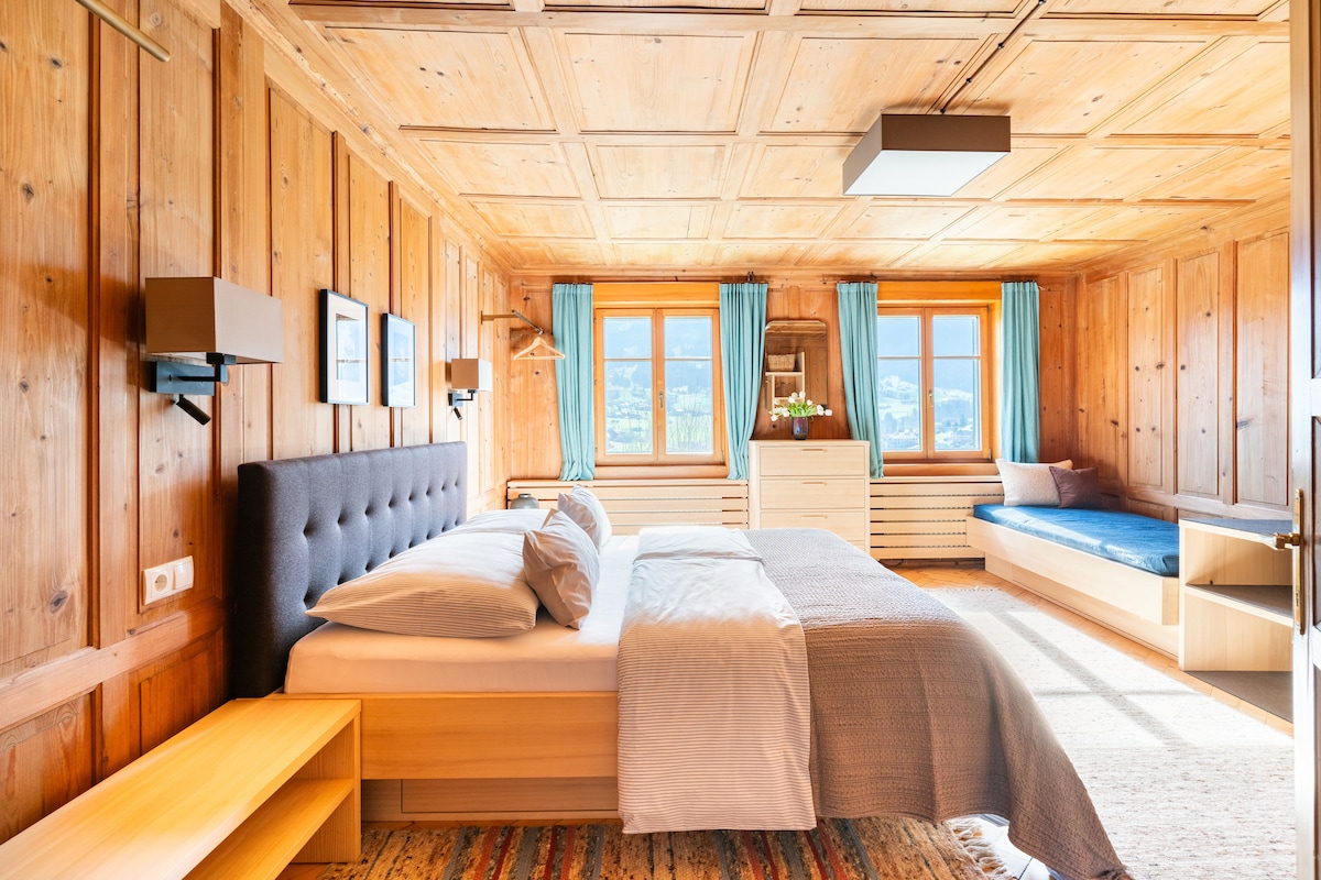 A spacious bedroom features traditional wood paneling and a coffered ceiling. A king-size bed is paired with a day bed against the wall. Large windows draped with blue curtains illuminate the room, while minimal furniture includes bedside tables and a dresser.