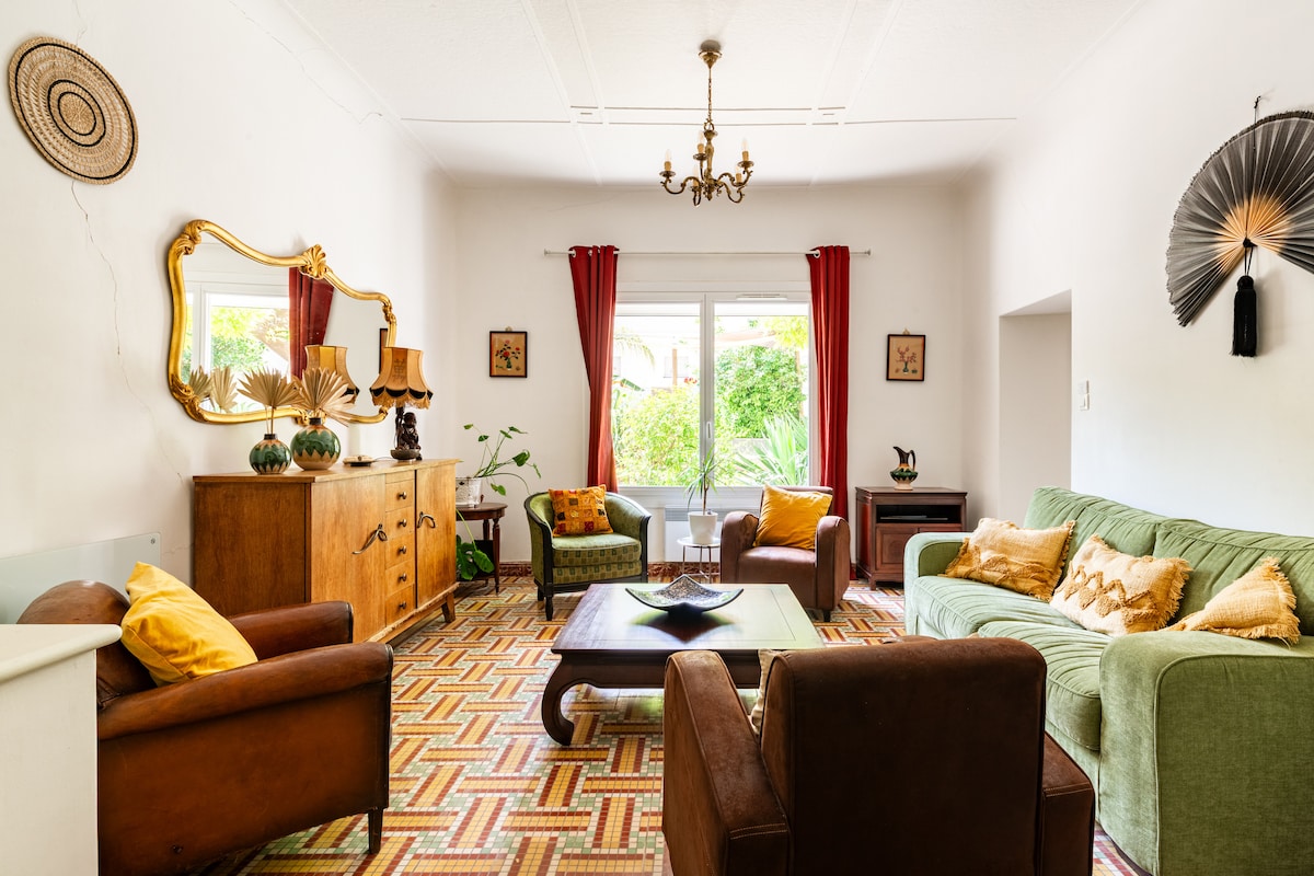 A spacious living room features a mixture of vintage and modern decor, including a green sofa and brown armchairs. A decorative mirror hangs above a wooden console table. Natural light flows through large windows, accompanied by warm curtains, while potted plants add a refreshing touch.