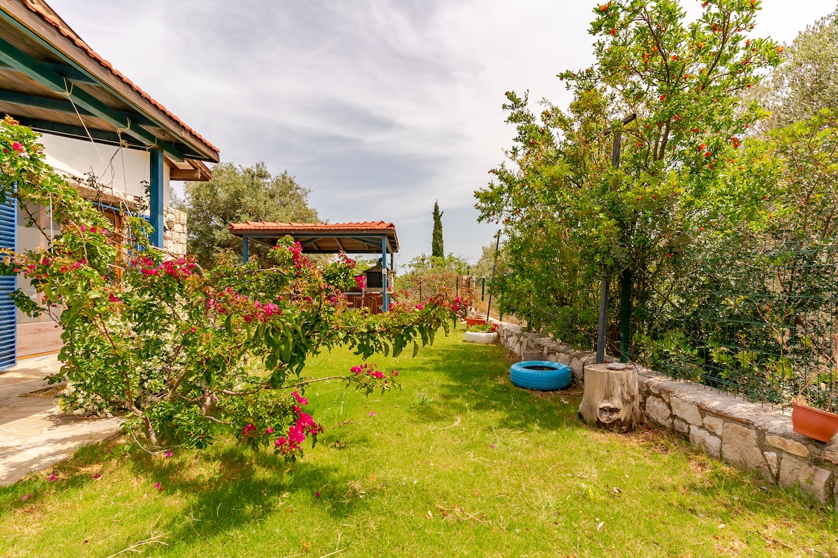 A well-maintained garden area is featured, with lush green grass interspersed with flowering plants. A shaded seating area is visible under a wooden structure, along with trees providing additional foliage. Traces of colorful flowers add vibrancy to the peaceful outdoor space.