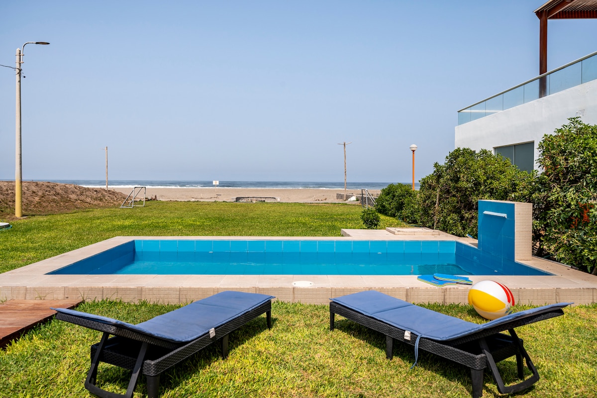 A private swimming pool is situated on a green lawn, surrounded by low shrubs. Two lounge chairs are positioned beside the pool, and a beach ball rests nearby. In the background, the sandy beach is visible, leading to the ocean under a clear sky.