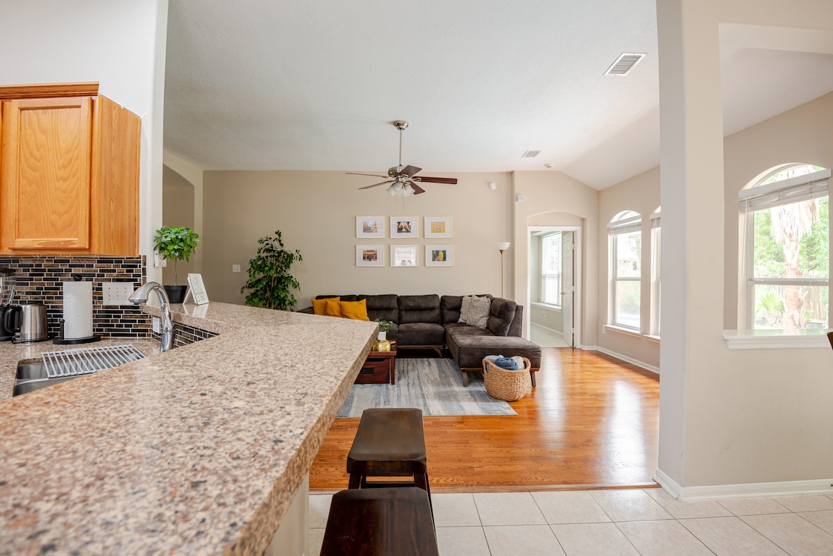 A spacious open-concept living area is highlighted by a large sectional sofa and a smooth granite countertop in the kitchen. Natural light enters through several windows, creating a warm ambiance. A small dining table sits in the corner, surrounded by soft decor.