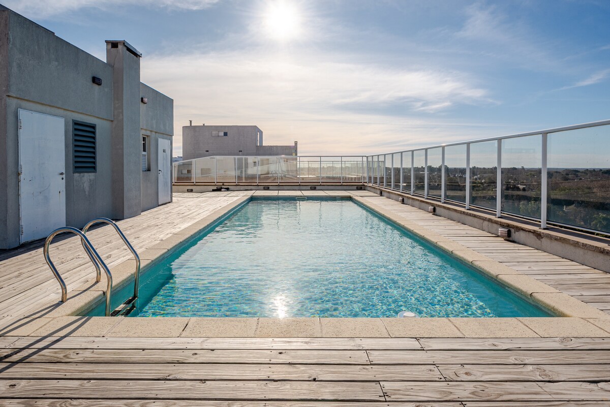 A rooftop swimming pool is surrounded by wooden decking, with a clear blue sky reflecting sunlight overhead. Glass railings provide unobstructed views of the surrounding area. The pool's water is calm and inviting, contributing to a serene atmosphere.