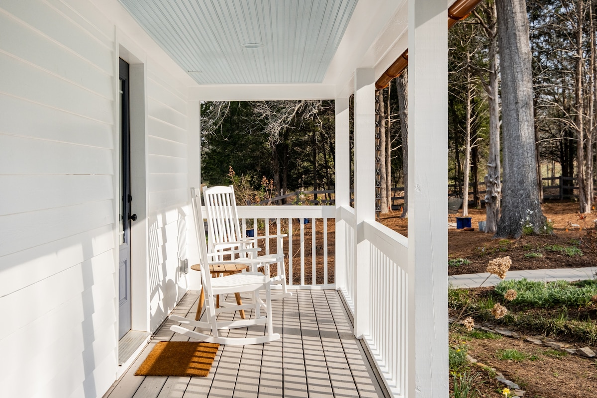 A serene porch area is showcased, featuring a white rocking chair on a light wood floor. Natural light filters through the porch's open layout, highlighting the surrounding trees and the gently manicured landscape.