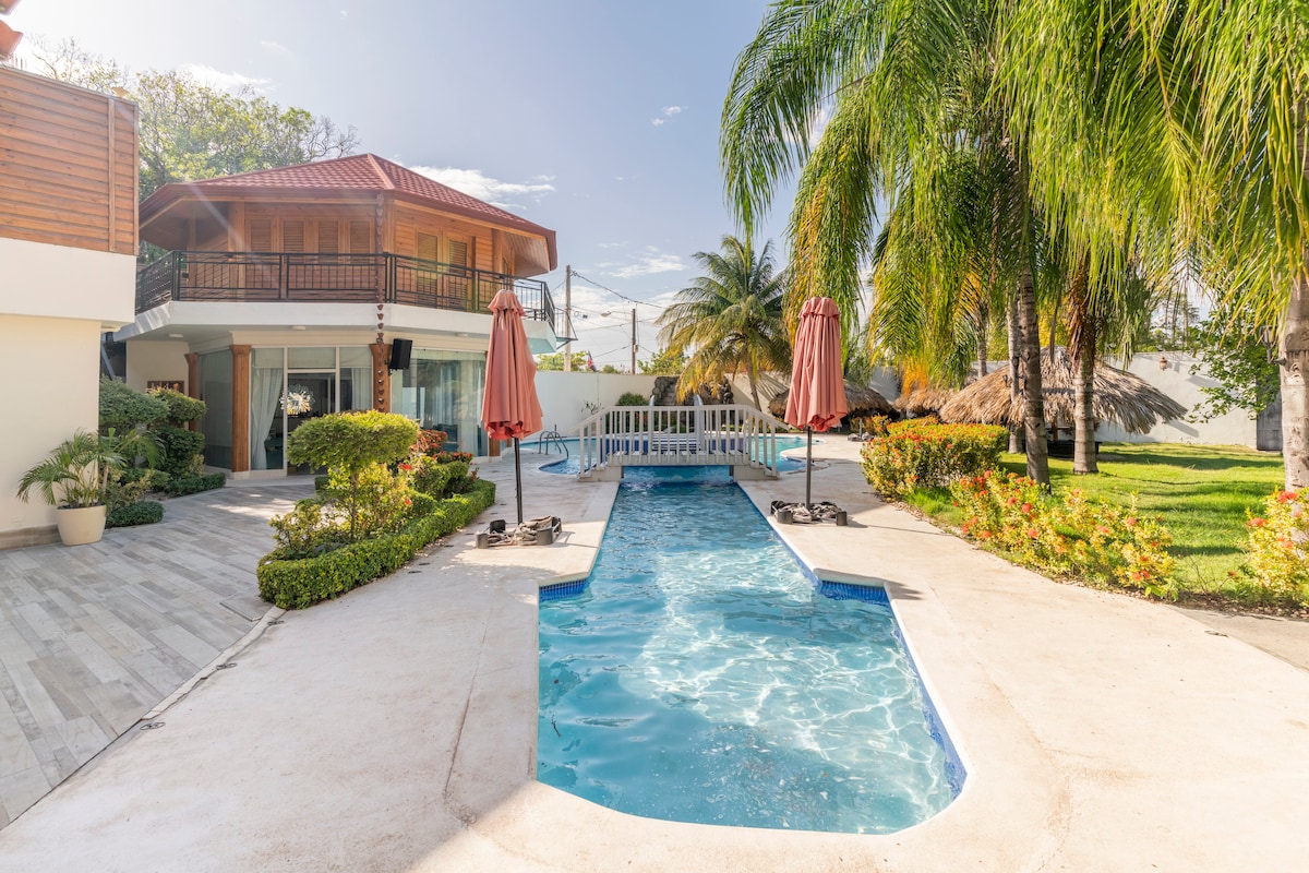 A private pool area features a cool, shimmering blue shape surrounded by lounge chairs and palm trees. The villa is visible in the background, showcasing a two-story structure with a red-tiled roof, offering a harmonious blend of indoor and outdoor spaces.