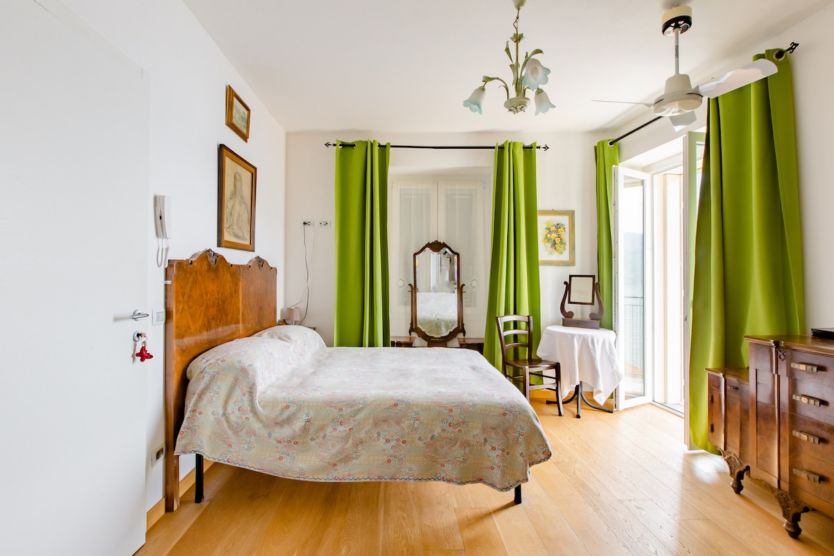A bright room features a spacious bed with a patterned coverlet. Green curtains frame the windows, allowing natural light to filter in. A vintage mirror and a small table are positioned across from the bed, enhancing the elegant decor. A wooden dresser adds warmth to the space.