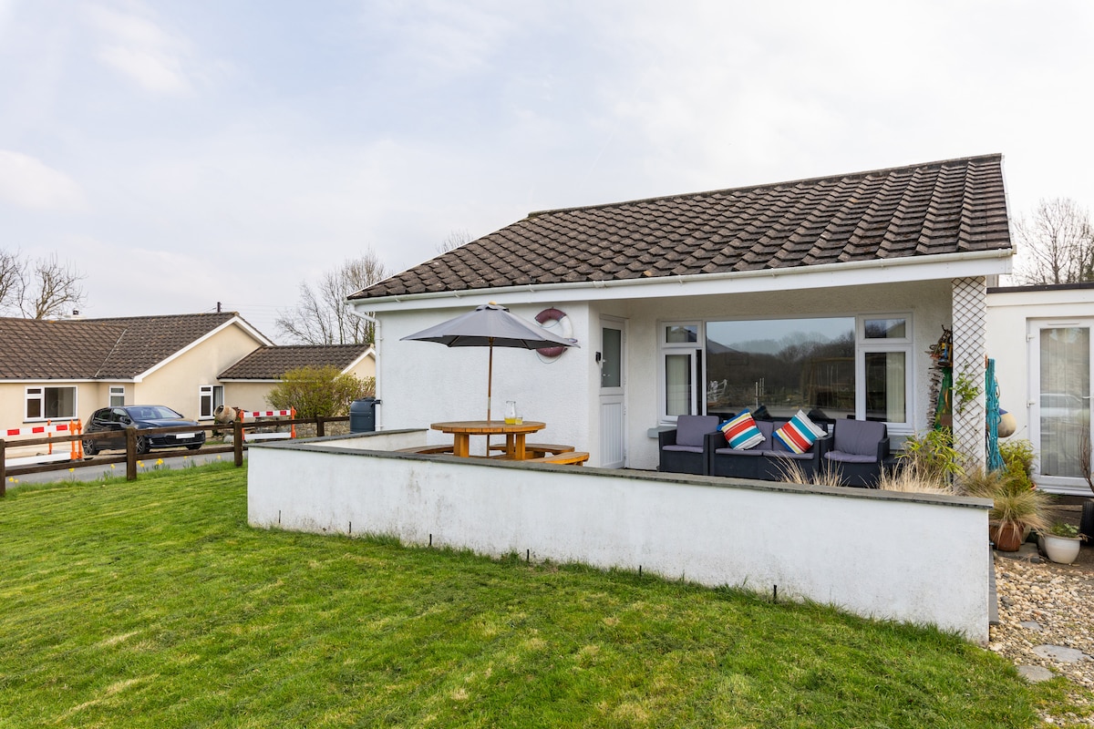 A family bungalow is displayed with a spacious outdoor seating area featuring a wooden table and comfortable seating options. An umbrella provides shade, while the lawn is well-kept. The surrounding scenery includes other residences and greenery.