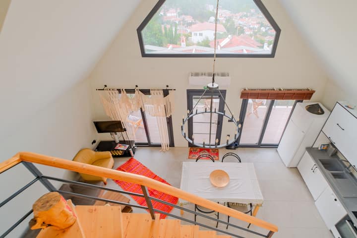 A cozy overhead view from the loft level, capturing the dining table, comfy sofa corner, entrance doors, and the bright, open-plan living space — perfect for your peaceful stay.