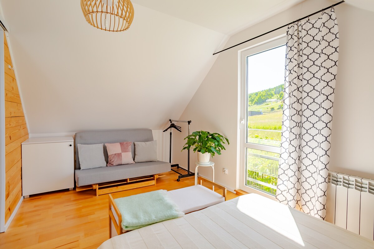 A well-lit bedroom features a spacious bed with light bedding. A cozy seating area includes a grey sofa. A large window allows natural light to fill the space and offers serene views of the surrounding hills. A potted plant adds a touch of greenery.