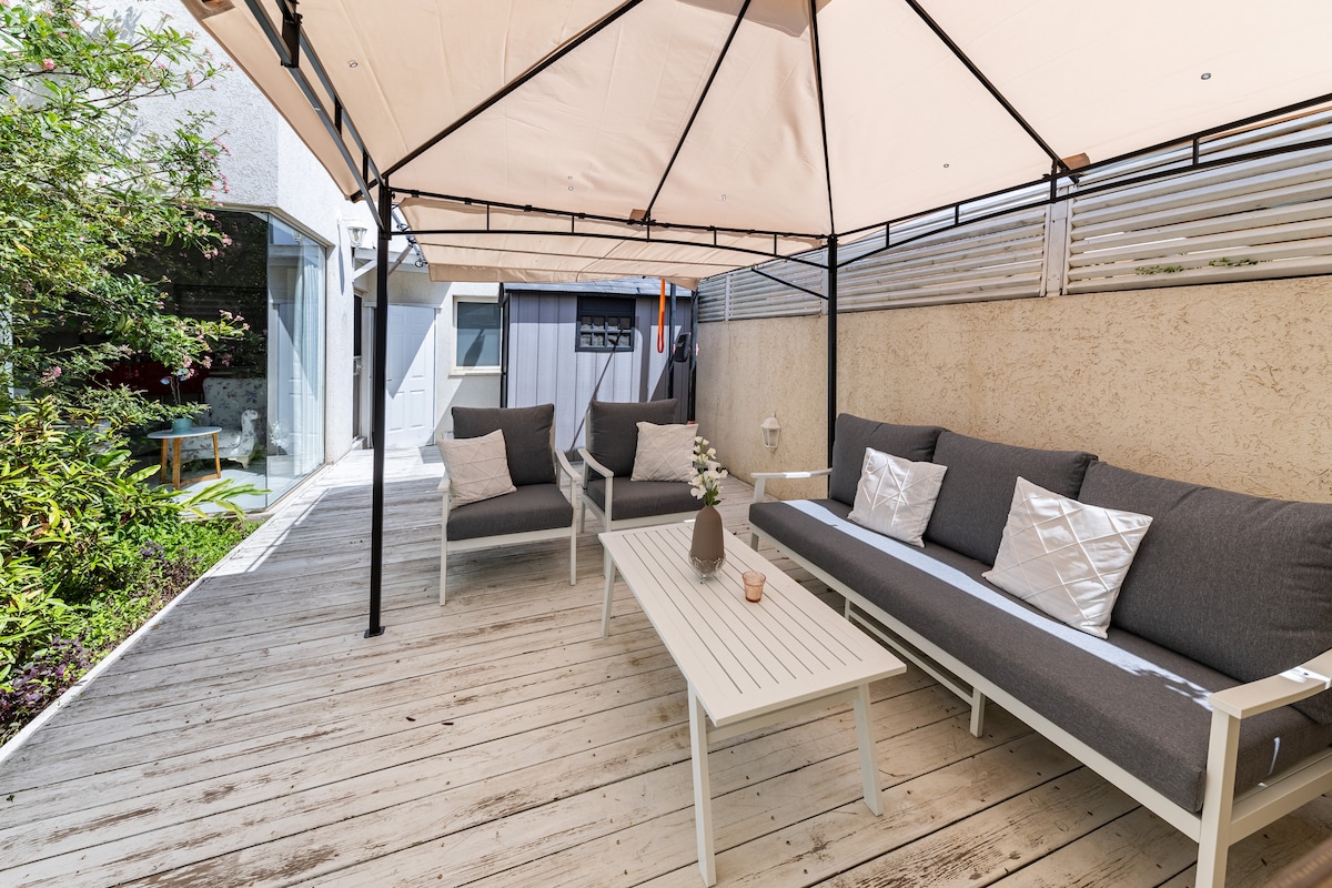 A shaded outdoor seating area is presented under a canopy, featuring a light wooden deck. Comfortable gray sofas are arranged around a low white table, complemented by decorative pillows. Lush greenery surrounds the space, providing a tranquil setting for relaxation.
