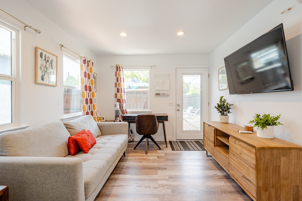 A bright living area features a light gray sofa with an accent pillow, paired with a wooden coffee table. A desk and chair are positioned near a door that opens to the outdoor space. Natural light fills the room through two windows adorned with playful patterned curtains.