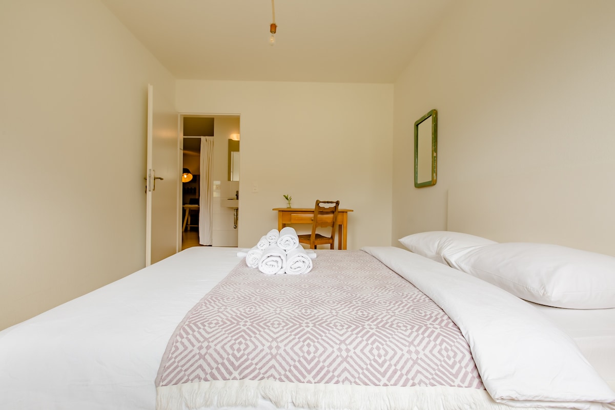 A bright sleeping area features a spacious bed topped with a patterned coverlet and neatly arranged towels. A wooden table with a chair is positioned nearby, and a doorway leads to a bathroom space, creating an open and inviting layout.