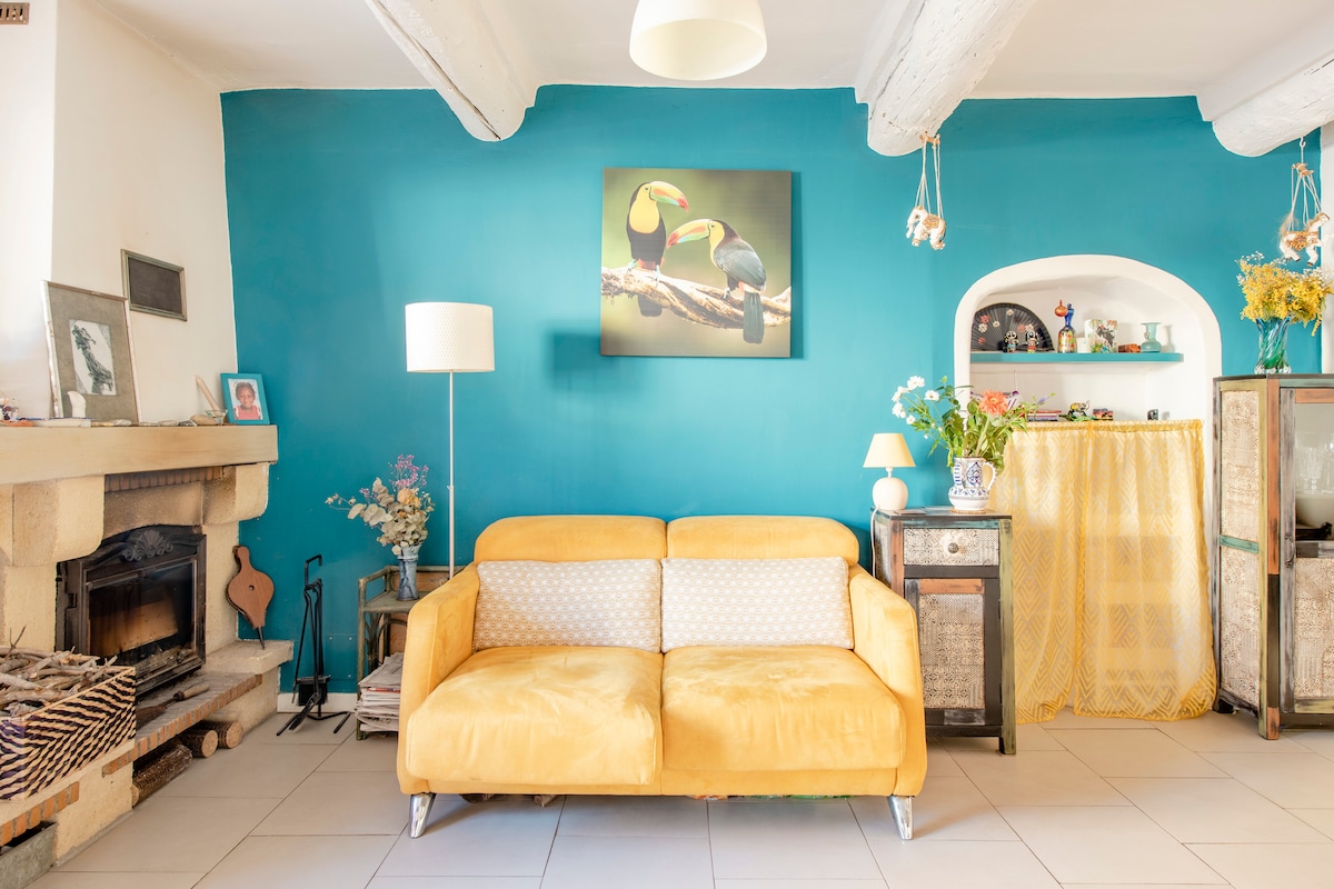 A vibrant living space is highlighted by a yellow sofa adorned with cushions. A blue accent wall features a framed painting of toucans. A light fixture hangs from the ceiling, and a stone fireplace adds character to the room. Decorative items and flowers are arranged throughout.