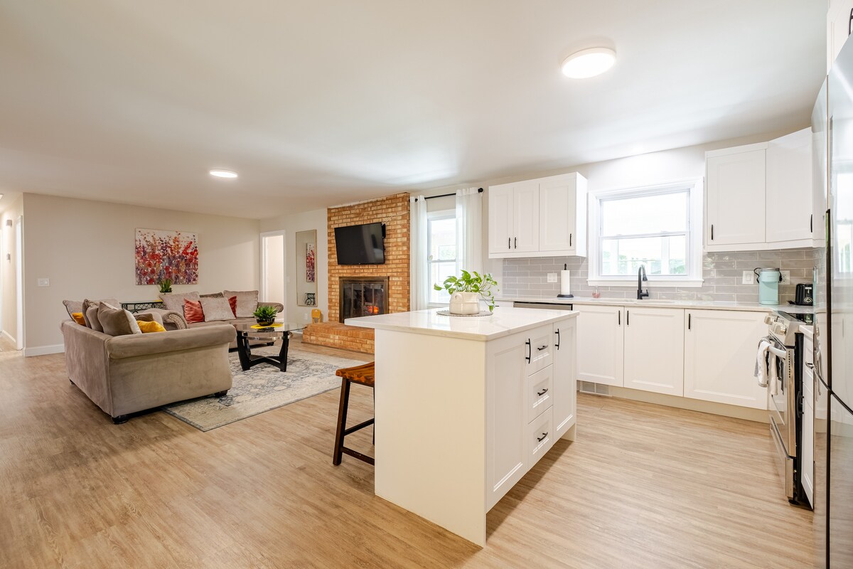 An open concept living space features a modern kitchen with white cabinetry, a large island with seating, and stainless steel appliances. A cozy sitting area is visible with a neutral-toned sofa and colorful accent pillows, while a warm brick fireplace adds a touch of comfort.