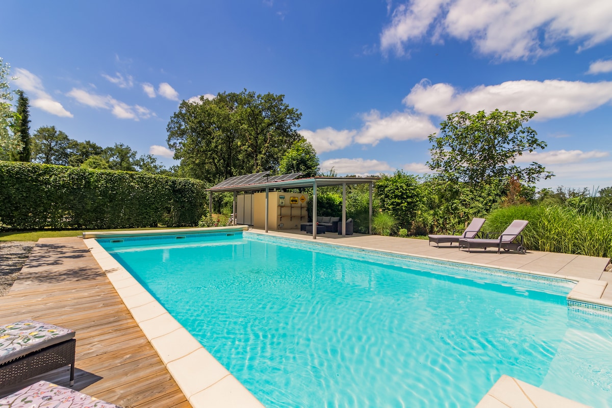 A rectangular swimming pool is set against a clear blue sky, surrounded by a spacious deck. Lounge chairs are positioned on one side, with greenery providing privacy along the perimeter. A covered seating area is visible in the background, enhancing the outdoor space.