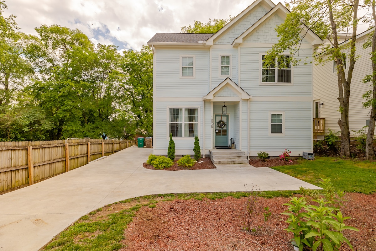 A newly constructed two-story home is displayed, featuring light-colored siding and a welcoming entryway. A neatly landscaped front yard includes low shrubs and flower beds, while a spacious driveway curves towards the left. Tall trees provide a natural backdrop, enhancing the serene setting.