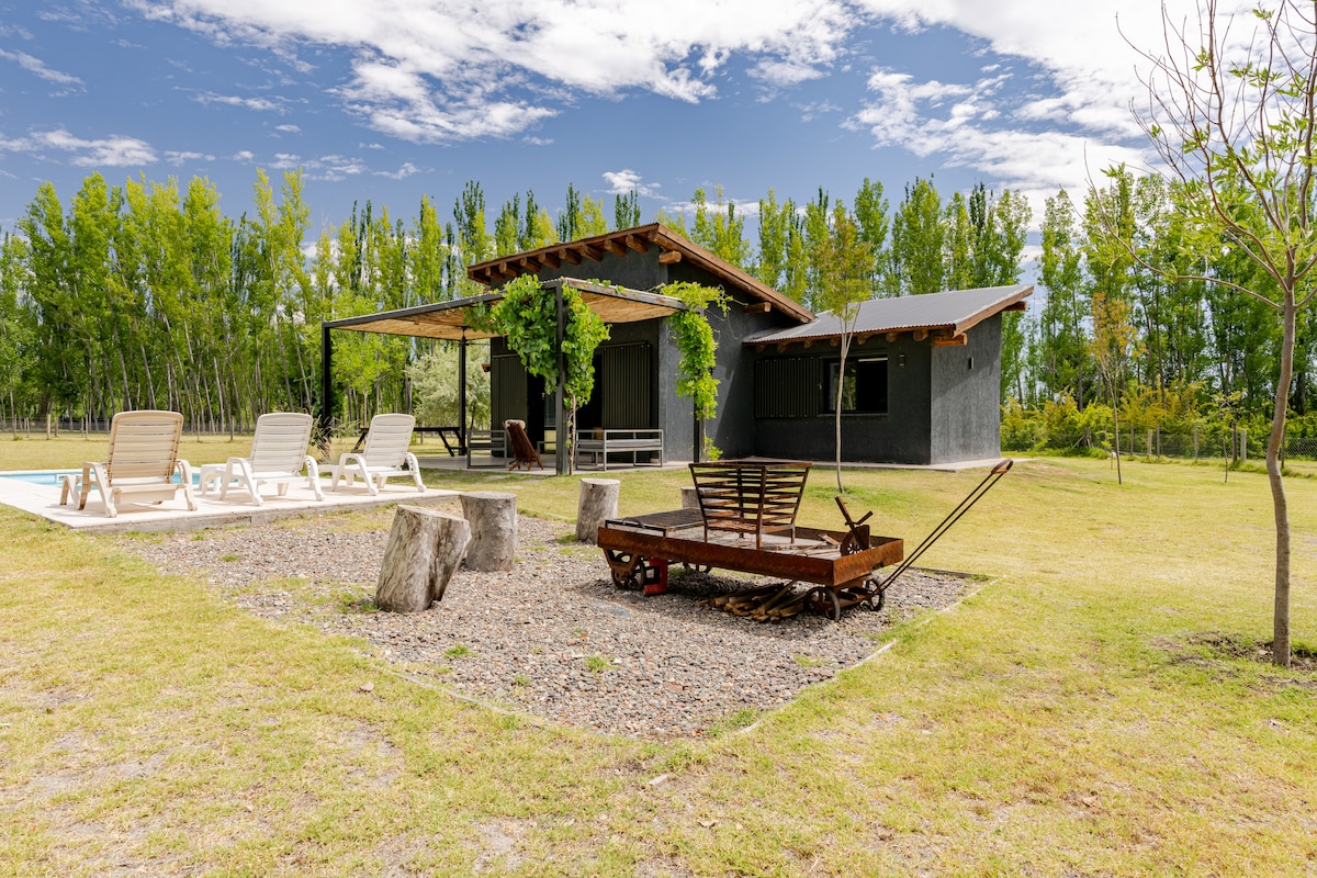 A spacious outdoor area features a well-maintained lawn surrounded by tall trees and natural greenery. A rustic cart is positioned prominently, while lounge chairs are arranged elegantly near the pool. A covered patio area provides shaded seating, complementing the serene setting.